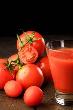 Yanında domates suyu olan cam bir bardak, bir domates dalı uzanıyor. Ahşap bir masanın üzerinde bir bardak domates suyu ve domates var. Siyah bir arka planda yatıyor..