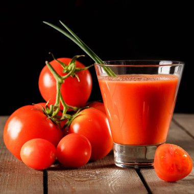 Yanında domates suyu olan cam bir bardak, bir domates dalı uzanıyor. Ahşap bir masanın üzerinde bir bardak domates suyu ve domates var. Siyah bir arka planda yatıyor..