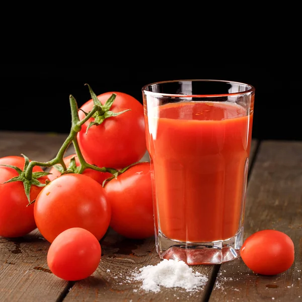 Yanında domates suyu olan cam bir bardak, bir domates dalı uzanıyor. Ahşap bir masanın üzerinde bir bardak domates suyu ve domates var. Siyah bir arka planda yatıyor..