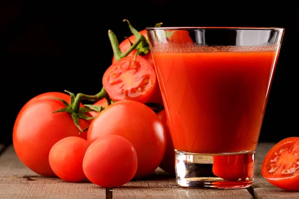 Yanında domates suyu olan cam bir bardak, bir domates dalı uzanıyor. Ahşap bir masanın üzerinde bir bardak domates suyu ve domates var. Siyah bir arka planda yatıyor..