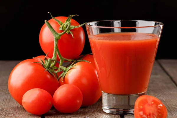 Yanında domates suyu olan cam bir bardak, bir domates dalı uzanıyor. Ahşap bir masanın üzerinde bir bardak domates suyu ve domates var. Siyah bir arka planda yatıyor..