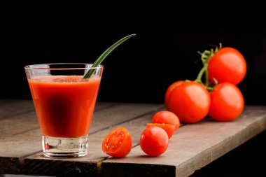 Yanında domates suyu olan cam bir bardak, bir domates dalı uzanıyor. Ahşap bir masanın üzerinde bir bardak domates suyu ve domates var. Siyah bir arka planda yatıyor..
