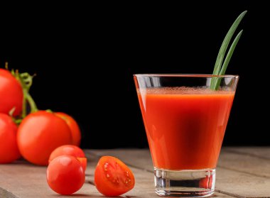 Yanında domates suyu olan cam bir bardak, bir domates dalı uzanıyor. Ahşap bir masanın üzerinde bir bardak domates suyu ve domates var. Siyah bir arka planda yatıyor..