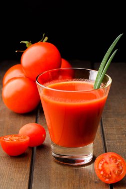 Yanında domates suyu olan cam bir bardak, bir domates dalı uzanıyor. Ahşap bir masanın üzerinde bir bardak domates suyu ve domates var. Siyah bir arka planda yatıyor..