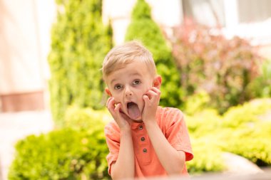 Beş yaşında bir çocuk surat yapıyor. Beş yaşında sarışın bir çocuk, güneşli bir havada sokakta dikilirken surat asıyor.