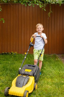 Sarışın bir çocuk sarı bir çim biçme makinesiyle bahçedeki çimleri biçiyor. Güneşli bir yaz gününde, altı yaşında bir çocuk sarı bir çimlerin çimlerini biçiyor..