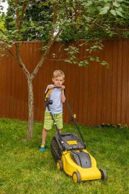 Sarışın bir çocuk sarı bir çim biçme makinesiyle bahçedeki çimleri biçiyor. Güneşli bir yaz gününde, altı yaşında bir çocuk sarı bir çimlerin çimlerini biçiyor..