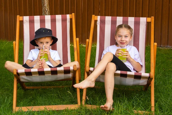On a sunny summer day, a girl and a boy of six years old (of European appearance) are relaxing in the garden on deck chairs and drinking a green drink.