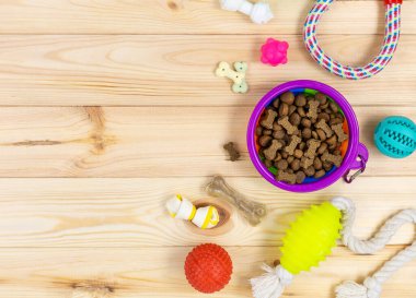 Farklı renkte hayvan bakım aksesuarları: menekşe kasesi, kemikler, toplar, atıştırmalıklar, doğal ahşap arka planda oyuncaklar. Köpekler için lastik ve tekstil aksesuarlar. Üst manzara, düz yatış. Boşluğu kopyala