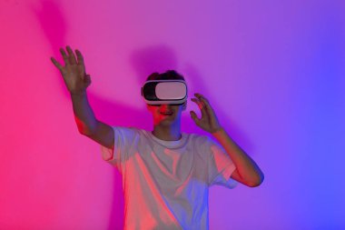 Modern VR kulaklık takarken gülümseyen beyaz tişörtlü sevimli bir genç pembe mor arka planda duruyor. Modern hobi, çocuklar ve gençler için video oyunları oynamak.