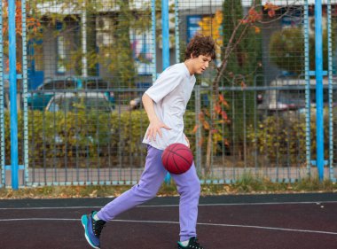 Dışarıda basketbol oynayan beyaz tişörtlü tatlı bir genç. Top öğrenme yeteneği olan genç bir çocuk şehir sahasında top sürüyor ve ateş ediyor. Çocuklar için hobi, aktif yaşam tarzı
