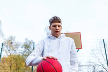Cute teenager in white hoodie playing basketball. Young boy with red ball learning dribble and shooting on the city court. Hobby for kids, active lifestyle