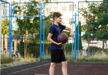 Mavi tişörtlü sevimli gülümseyen çocuk şehir parkında basketbol oynuyor. Turuncu bir topla açık hava oyununun tadını çıkaran aktif bir genç. Hobi, aktif yaşam tarzı, çocuklar için spor, gençler