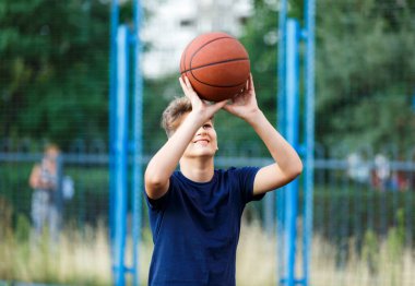 Mavi tişörtlü sevimli gülümseyen çocuk şehir parkında basketbol oynuyor. Turuncu bir topla açık hava oyununun tadını çıkaran aktif bir genç. Hobi, aktif yaşam tarzı, çocuklar için spor, gençler