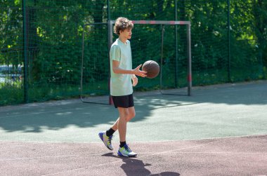 Basketbol oynayan genç, gülümseyen, neşeli. Basketbol topu olan zinde bir çocuk. Sahada top süren çocuk.