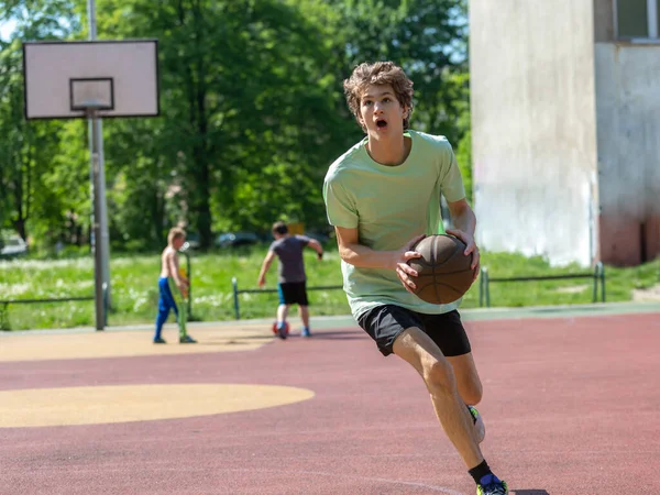 Teenager playing basketball smiling, cheerful. Fit boy with basketball ball outdoors. Kid dribbling the ball at the court