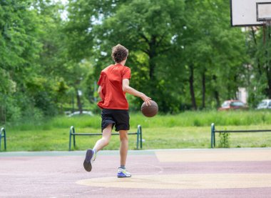 Teenager running in the stadium. Cute young teenager in red t shirt with a ball plays basketball on court. Sports, hobby, active lifestyle for boys