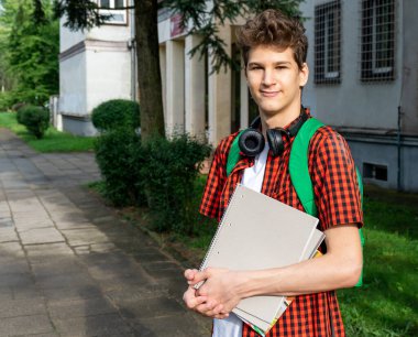 Cute teenager in red shirt, headphones, backpack holding a notebook in front of his school. Back to school background.