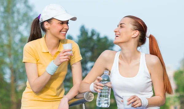 Two smiling girl friends in sports clothing drinking water, — Stock Photo, Image