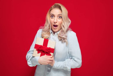 Elinde kırmızı arka planda izole edilmiş kurdeleli bir hediye kutusu olan sürpriz bir kız. Bayramlarda hediye getiren genç bir kadın, Noel 'de. Stüdyo çekimi