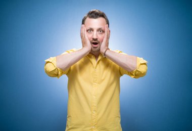 Mavi arka plan üzerinde elleri olan, sarı gömlekli, şaşkın bir adam. Şok olmuş bir adam. Stüdyo çekimi