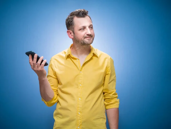 打电话的男人很生气 对着蓝色的背景 穿着黄色的衬衫 做了个鬼脸 讨厌的家伙说话 智能手机上的无聊和无趣 工作室拍摄 — 图库照片
