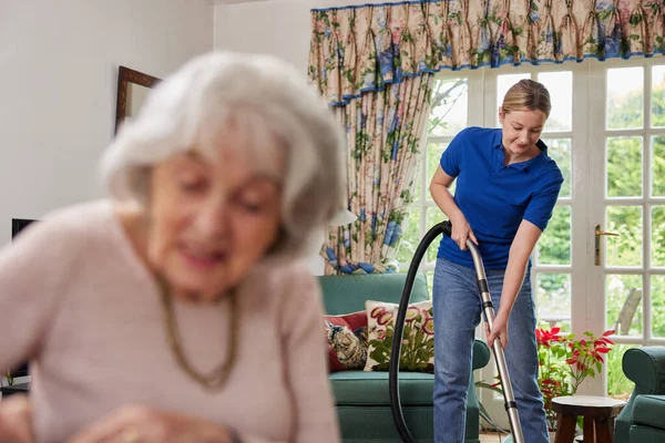 Vrouwelijke Thuishulp Schoonmaken Huis Met Stofzuiger Terwijl Senior Vrouw Leest — Stockfoto