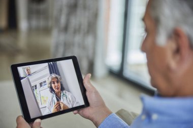 Evde dijital tablet kullanan bir adam doktorla uzaktan danışmanlık yapıyor.