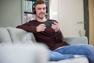 Evde kablosuz kulaklık takan adam cep telefonundan müzik ya da film izliyor.
