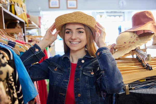 Young Woman Buying Used Sustainable Clothes From Second Hand Charity Shop Trying On Hat