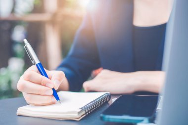 Kalemle not defterine el yazısı yazan bir kadın ve ofisinde cep telefonu tutuyor..