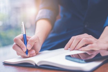 Bir kadın kalemle bir not defterine el yazısı yazıyor ve elinde de bir cep telefonu var. Web pankartı..