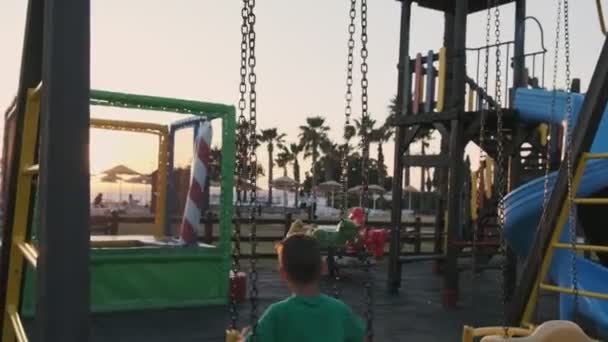 Boy monta em balanços, em câmera lenta em um playgound em uma ilha tropical ao pôr do sol — Vídeo de Stock