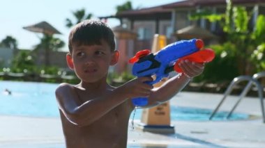 Tropik bir tatil köyünde halka açık bir havuzda oyuncak su tabancası ateşleyen küçük bir çocuğun yakın çekimi.