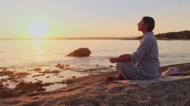Deniz kenarında dinlenme egzersizi yapan genç bir kadın. Denizin üzerinde altın gün batımı yoga yapıyor. Kumsalda gün batımı güneşin önünde yavaş yavaş meditasyon yapıyor.