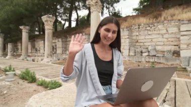 Genç bir kadın parkta oturmuş video görüşmesiyle iletişim kuruyor. Laptop bilgisayarına bakan bir kız, online konferans ofis sohbeti, sanal eğitim konsepti. Yavaş çekim