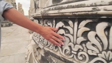 Eski tuğla duvara karşı ağır çekimde kayan bir kadın. Kadın eli, antik tapınağın sert yüzeyine dokunuyor.