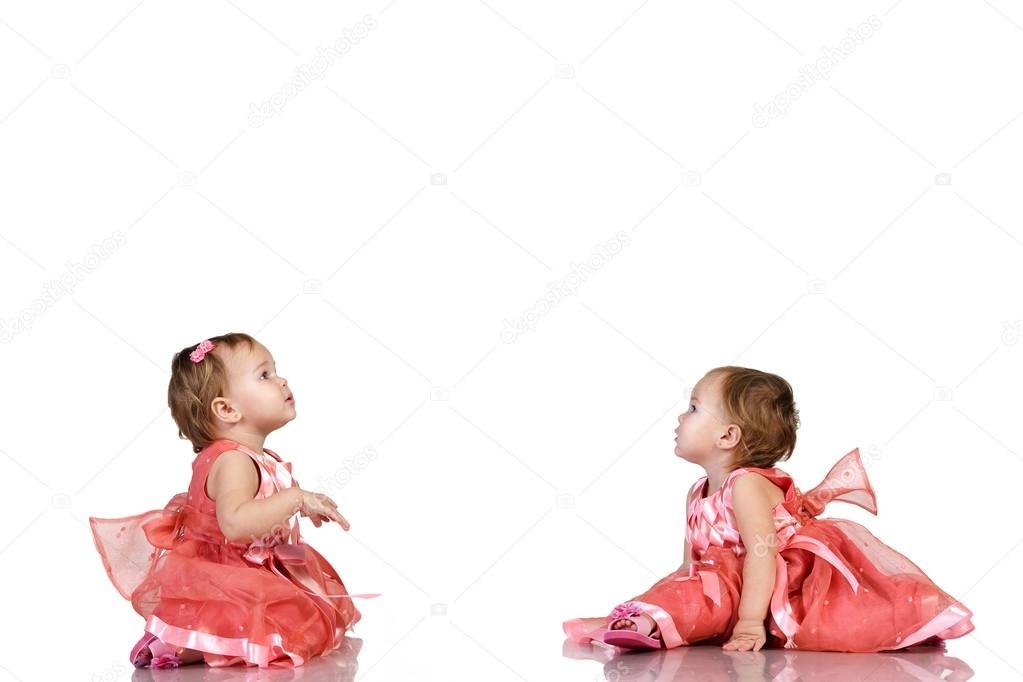 Identical twin baby girls in an elegant pink dresses looking carefully at something in their birthday. Place for your text or logo, isolated on white background.
