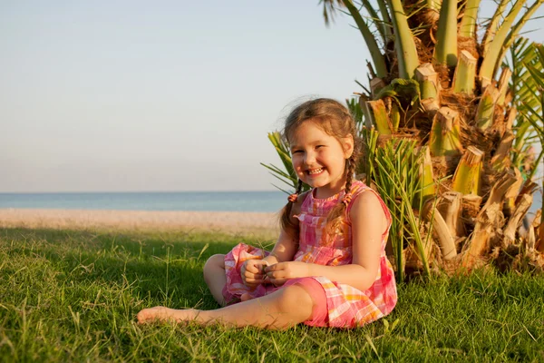 Kleines lächelndes Mädchen, das neben der Palme am Strand sitzt. — Stockfoto