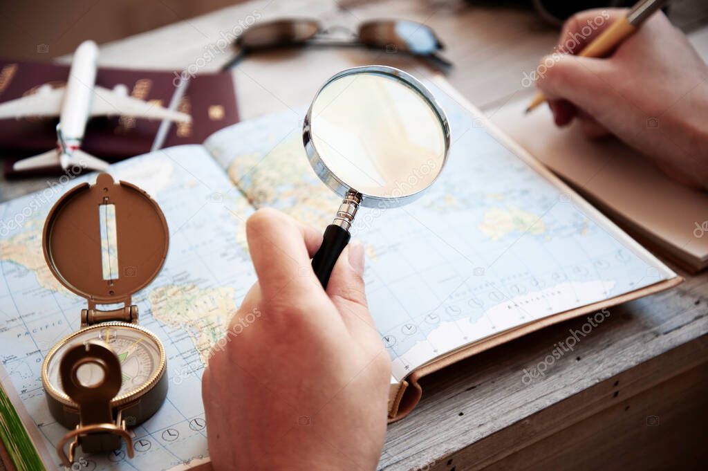 Male hand holding a magnifying glass for planning trip with world map on the table. Travel concept. Sunglasses, Passports, Paper notebook, Compass, camera, toy plane. The concept of preparing for the journey to meet adventures in the world.
