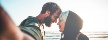 Gün batımında plajda selfie çeken aşık bir çiftle yatay afiş ya da başlık. Manyetik bakışlı bir kadın ve hipster erkek arkadaşı birbirlerinin gözlerinin içine bakarak selfie çekiyorlar.