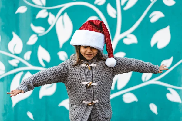 飛行機のようなオープンアームで飛ぶサンタクラスの帽子を身に着けている小さな女の子 面白い子供はクリスマスの時間に屋外の青い壁の背景に対して遊んで メリークリスマスと幸せな新年 — ストック写真