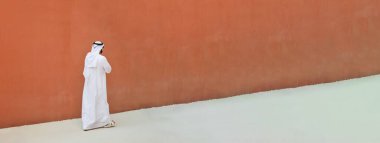 Arap kökenli, geleneksel emirlik elbiseli, turuncu duvarlı arka planda sokakta yürüyen Arap bir adamın yatay bayrağı ve Müslüman dini kavramı - metin için alan kopyala