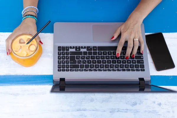 Female hands typing on laptop during remote working outdoors - Modern laptop on the blue wooden desk - Orange juice in a retro glass jar for healthy drink break - Remote working concept - Top view