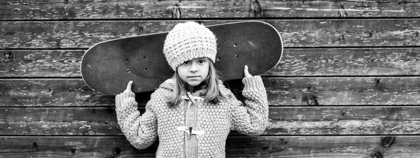 Horizontal banner or header with portrait of child girl skateboarder in wool winter clothes with skateboard on his shoulders looking at camera on wooden background - Black and white