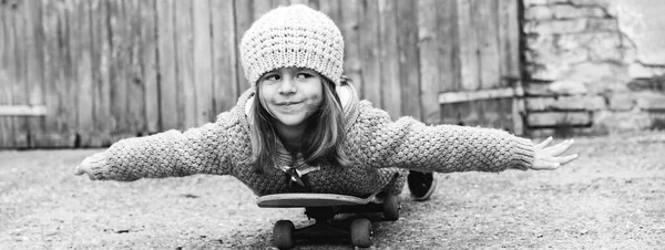 Horizontal banner or header with adorable child girl in winter wool clothes lying skateboard flies like a plane outdoors in backyard - Little girl playing with skateboard - Vintage black and white