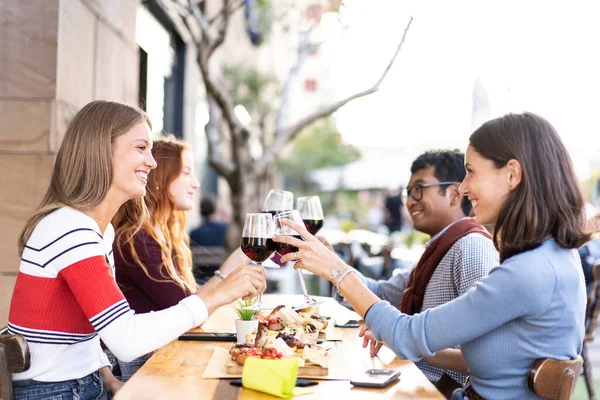 Multiracial friends toasting red wine at the outdoor pub in the city - Food and beverage lifestyle concept with happy people having fun together at open air pub - Bright filter with focus on glasses