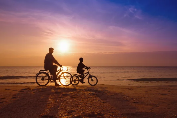 Vater und Sohn am Strand bei Sonnenuntergang, Familiensilhouette der Biker Stockbild