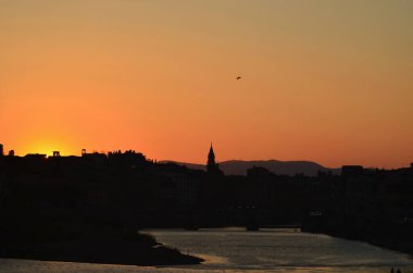 Floransa, İtalya 'da güneş batarken gökyüzü turuncuya döndü. Şehir siluet içinde, gökyüzünde çan kuleleri yükseliyor. Tepeler uzakta. Gökyüzünde tek bir kuş var..