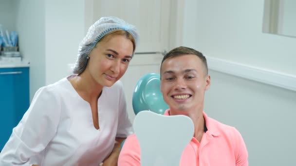 Σίγουρα όμορφος άντρας ασθενής της οδοντιατρικής κλινικής απολαμβάνει εκπληκτικό χαμόγελο μετά τη θεραπεία. Γυναίκα γιατρός και νεαρός άνδρας χαμογελούν κοιτάζοντας ο ένας τον άλλον. Στοματική υγιεινή στη σύγχρονη οδοντιατρική. Ο άνθρωπος κοιτά στον καθρέφτη — Αρχείο Βίντεο